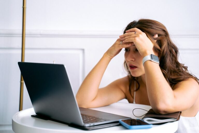 Woman feeling stressed while working on a laptop, using smartphone and smartwatch at a desk.
